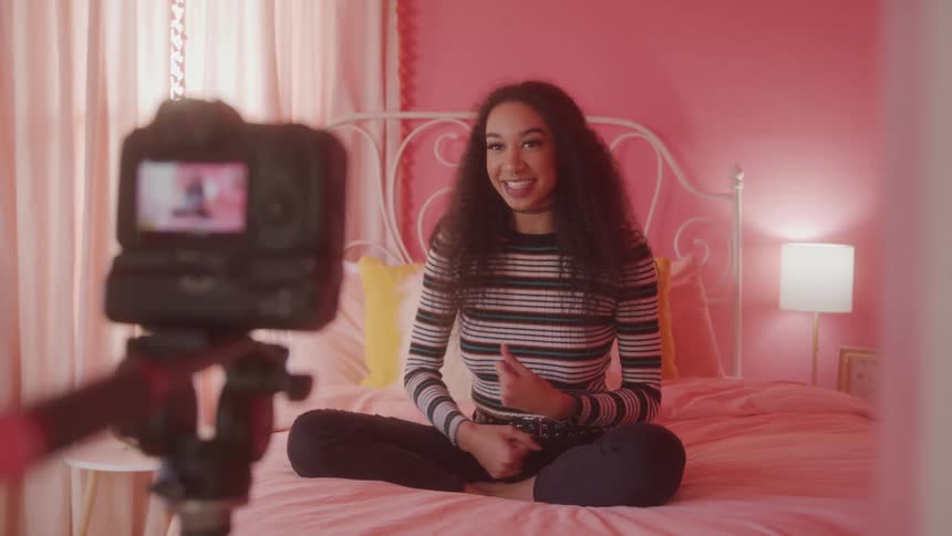 A woman sits crosslegged on a bed speaking enthusiastically to a camera