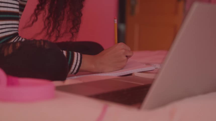 A woman writes in a book at a desk with a pencil with a laptop computer in front of her around midday