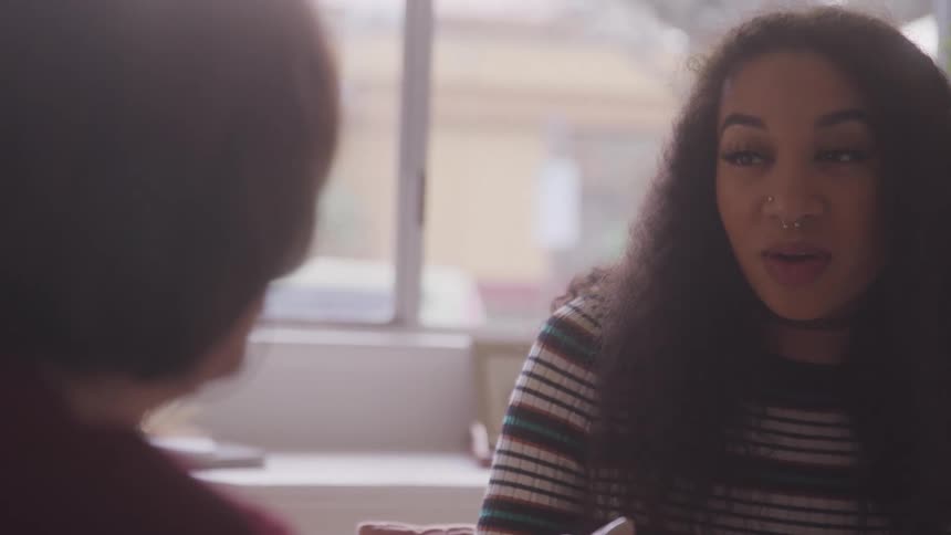 A young woman talks to another woman as they both sit at a coffee table indoors around midday