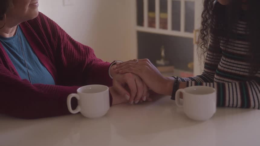 Wo people hold hands in a comforting gesture while sitting at a table with coffee mugs