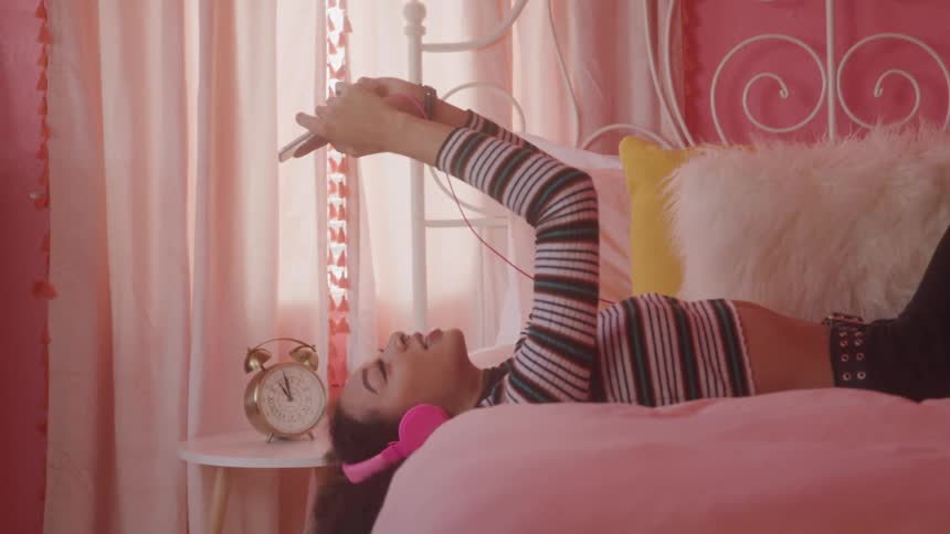 Girl lying on bed with headphones using phone in pinkthemed room