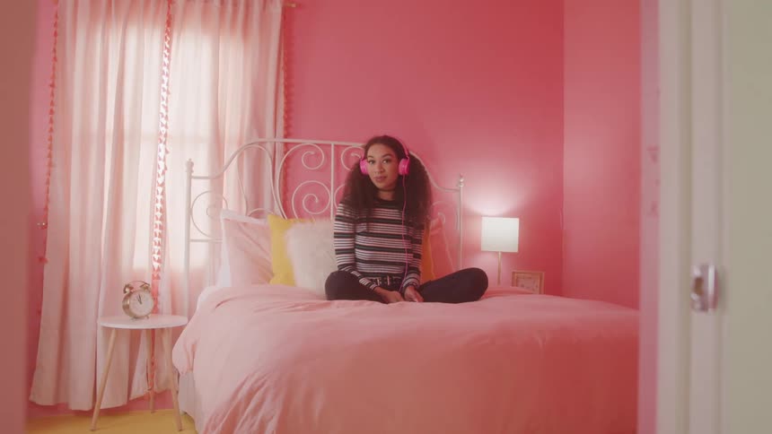 A young woman wearing pink headphones sits cross legged on a pink bed in a pink room