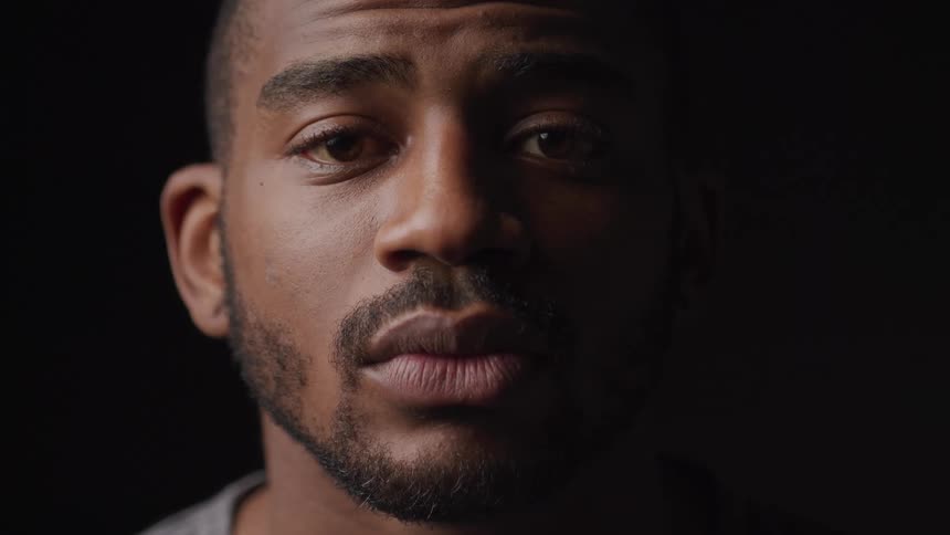 A young black man with a short beard and mustache sits in a dark room looking sad