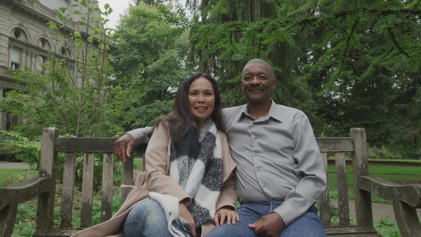 A smiling couple sits close together on a wooden bench in a green park