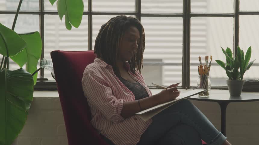 A young woman with dreadlocks sits comfortably in a red chair sketching in a notepad in a sunlit loft apartment