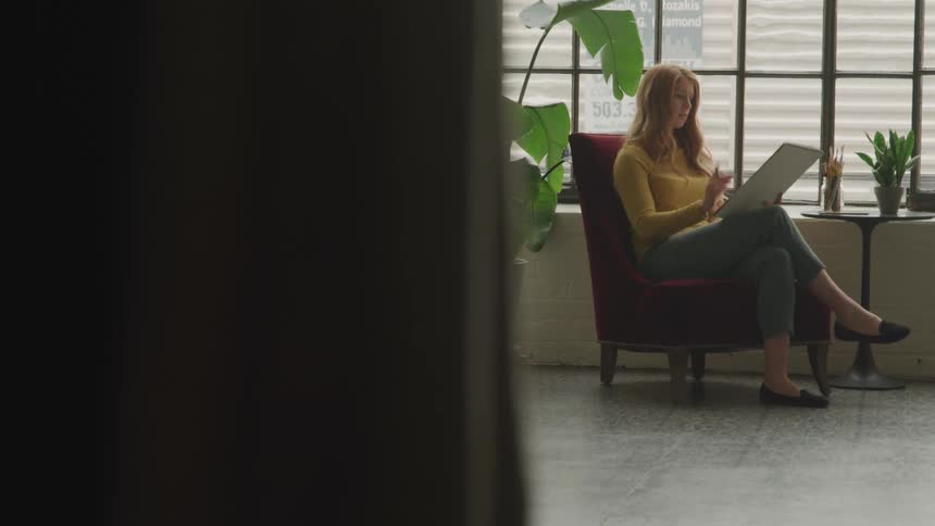 A woman sits on a red chair in a sunlit studio painting