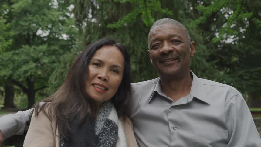 A couple sit on a bench side by side one wrapping an arm around the other near a forest