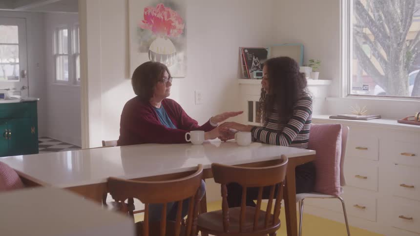 Two women sit at a kitchen table holding hands sharing a heartfelt moment