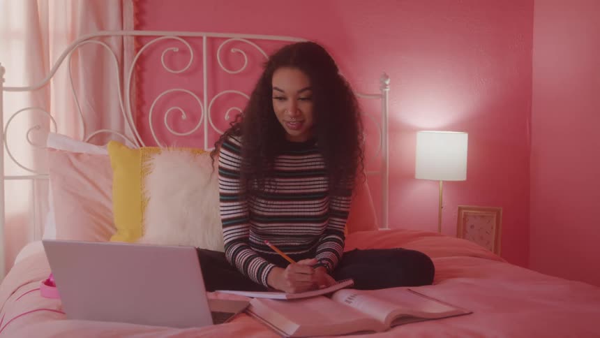 A young woman studies with books and a laptop while sitting on a bed in a pink bedroom