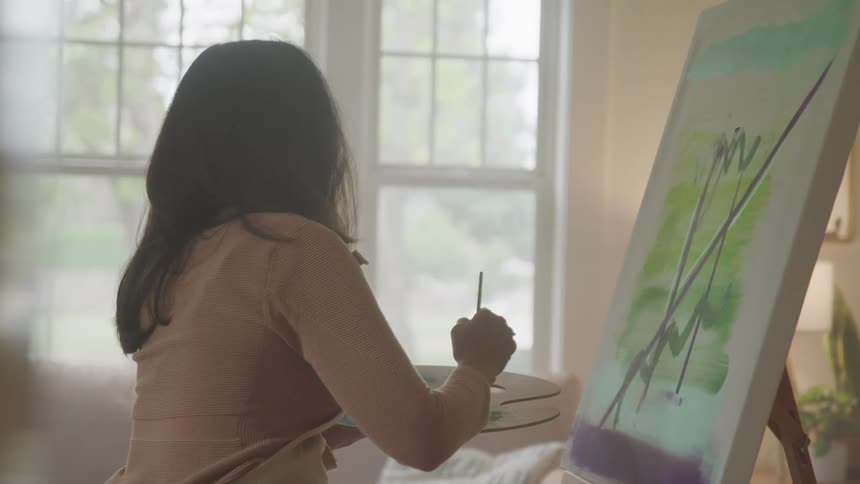 A woman sits by a window painting on a canvas in a softly lit room