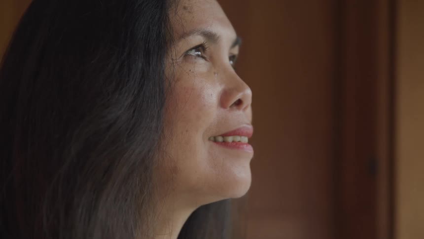 A woman with long black hair is indoors around midday with a smile on her face