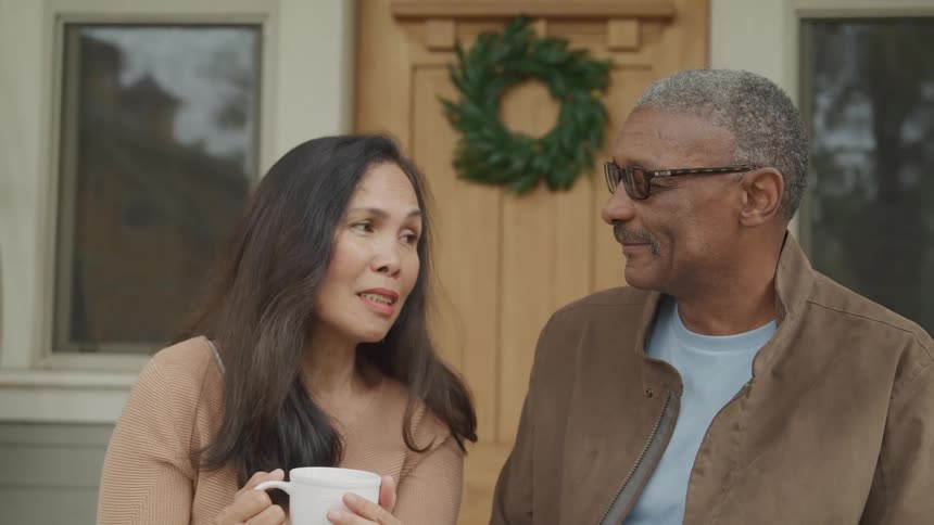 A middleaged couple sits on their front porch around midday engaged in a discussion
