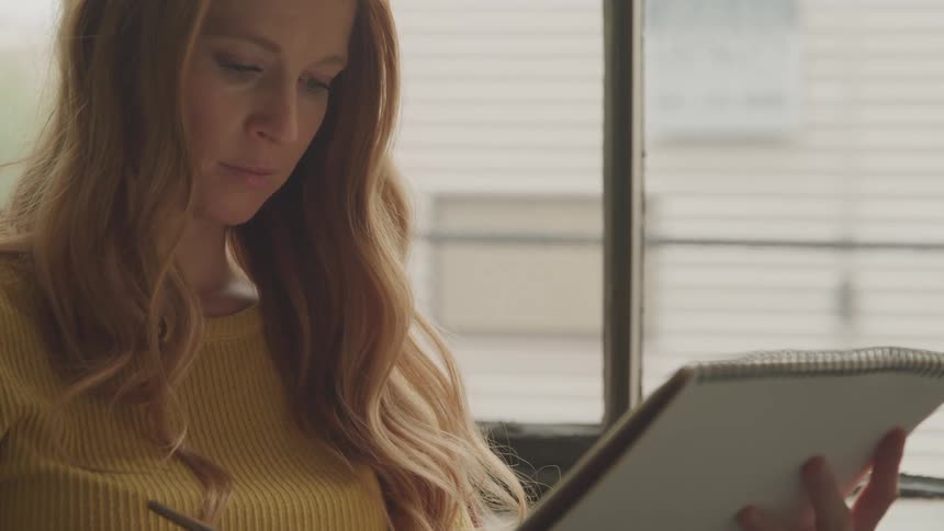 A woman is sitting in front of a window and sketching on paper