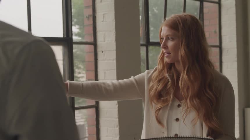 A young woman talks to another person pointing out the window while holding a pen and a book then writes afterward around midday