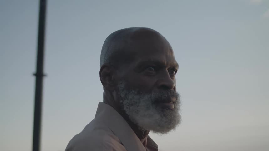 An older man stands by a calm riverside at dusk gazing thoughtfully into the distance