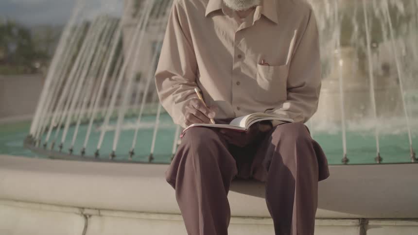 A fountain with an elderly black man sitting on its edge writing in a book around midday