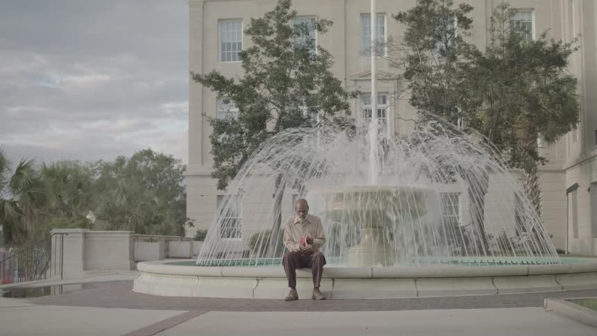 The man is writing in his notebook by the fountain