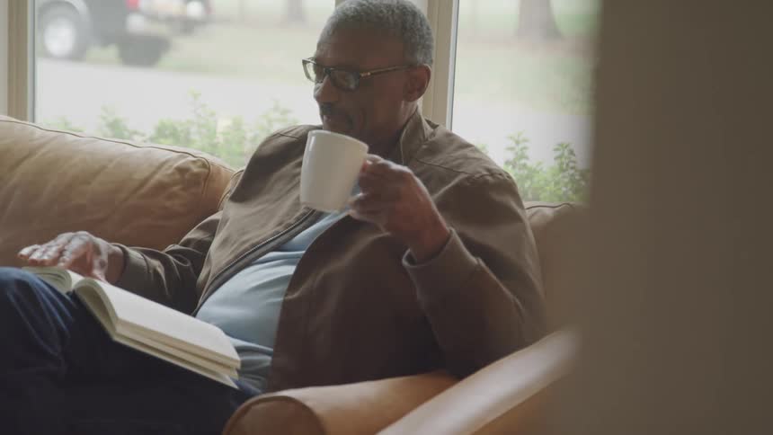 A man relaxes on a couch while reading a book and enjoying coffee by a large window