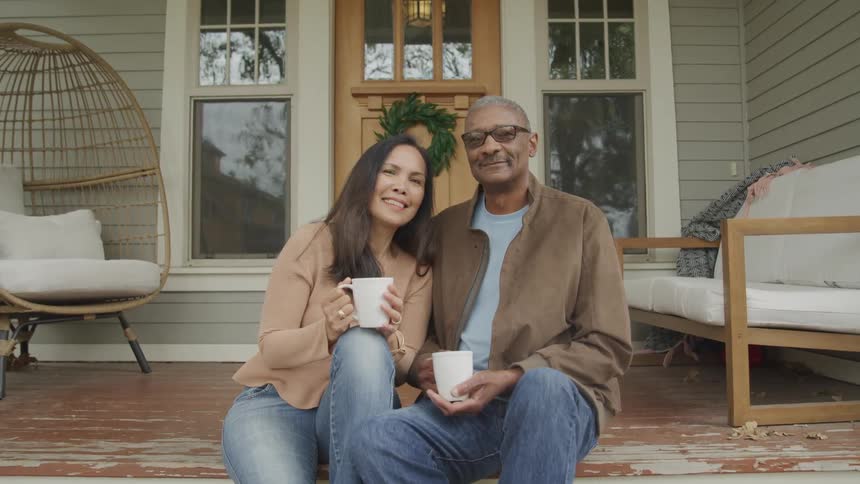 A couple consisting of a middleaged black man wearing glasses and a brown jacket and a woman both hold cups of coffee while sitting on their front porch around midday smiling