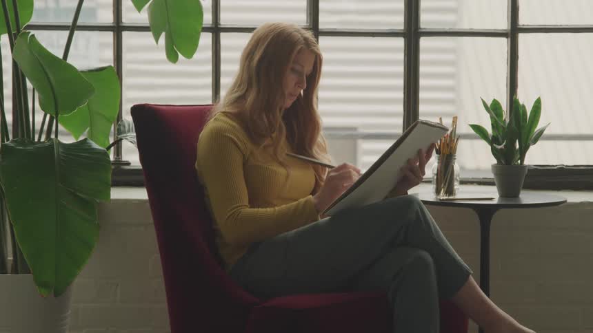 A woman sits by a window painting in a sketchbook with focus and calm