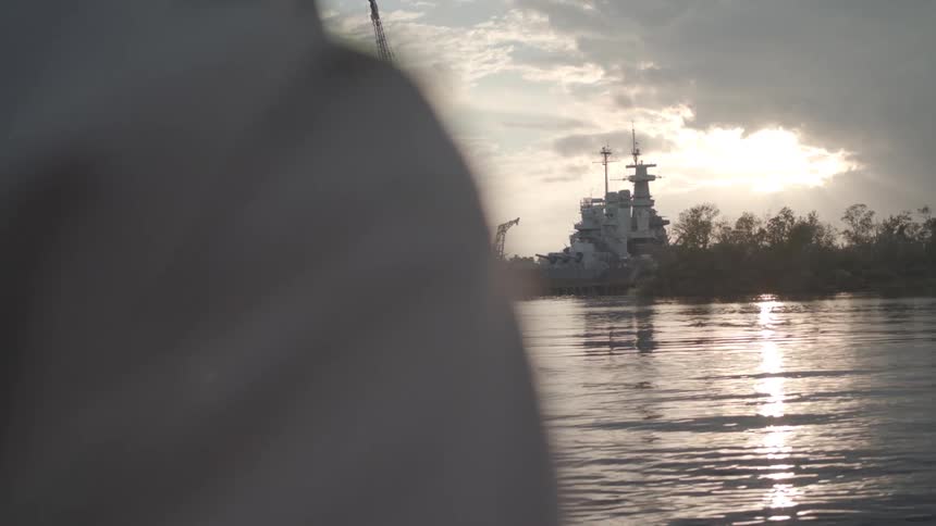 A large ship lies beside cranes and scaffolding at a harbor construction site viewed over a persons shoulder across calm water under a golden sunset