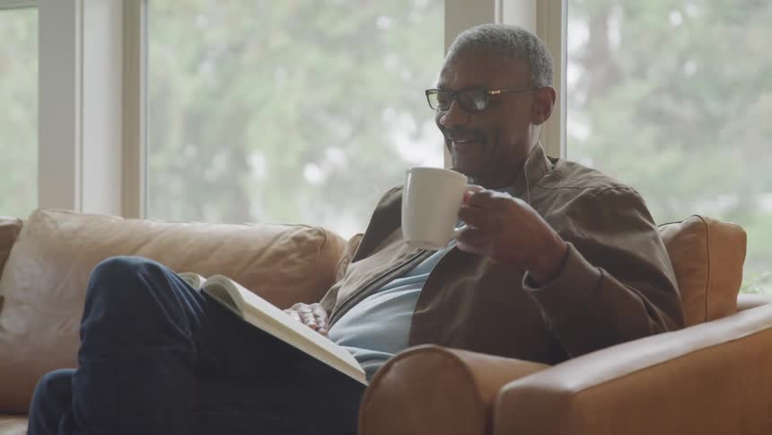 A man relaxes on a couch reading a book and enjoying a cup of coffee