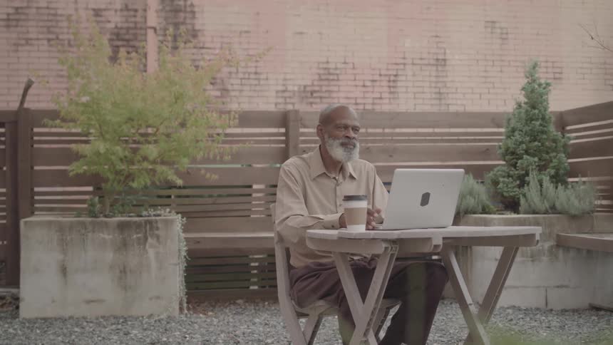 An elderly man works on a laptop at an outdoor table with coffee beside him