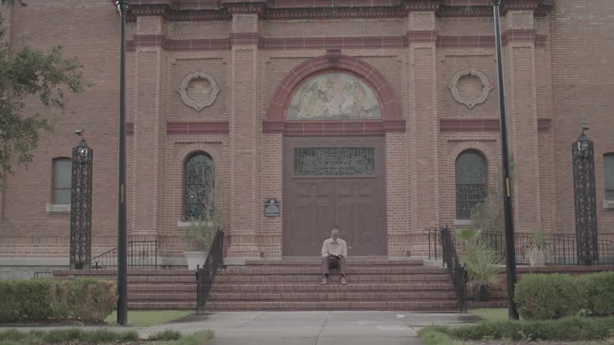 Person sits alone on steps in front of a brick building with wooden double doors and stained glass windows on the sides