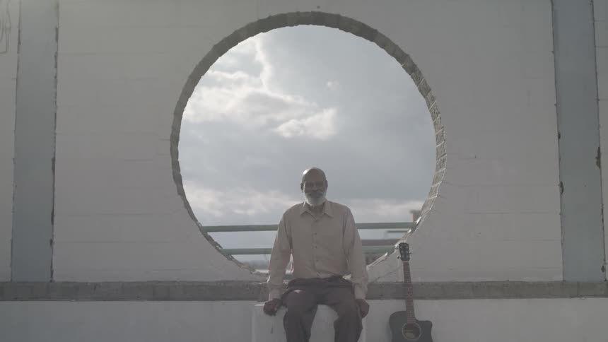 A man with a beard sits on a ledge with an acoustic guitar leaning against the wall framed by a large circular opening showing a cloudy sky