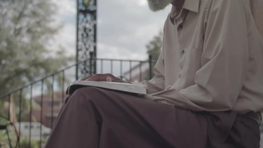 An elderly black man sits beside a building reading a book around midday