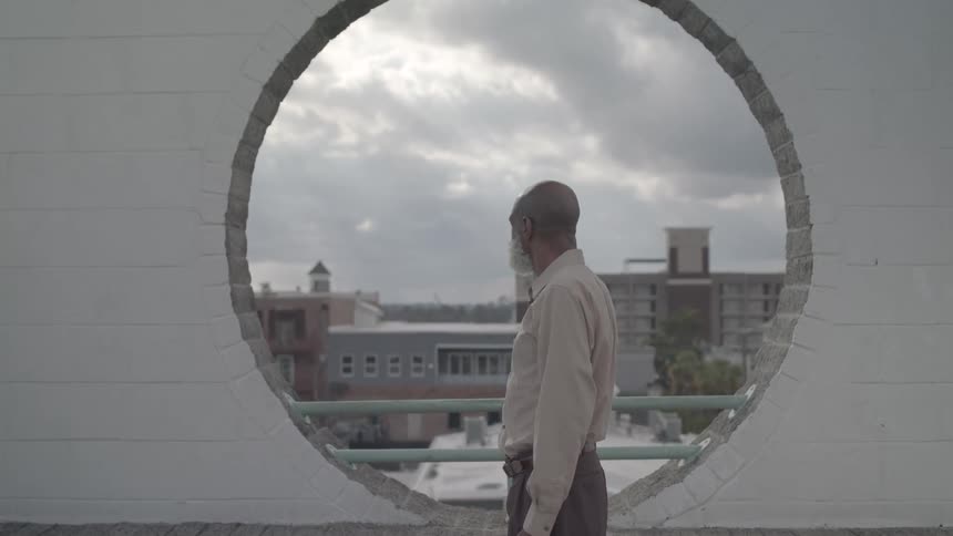 An elderly man stands before a circular wall opening with a cloudy city backdrop