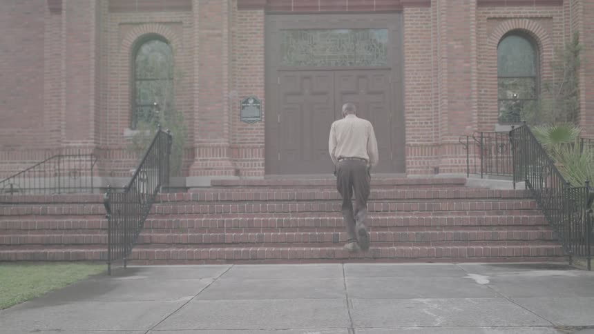 A person carrying a book sits on the steps of a large brick building with arched windows and a heavy wooden door