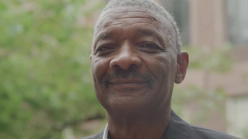 A man with a mustache smiles while looking directly at the viewer with out of focus foliage in the background