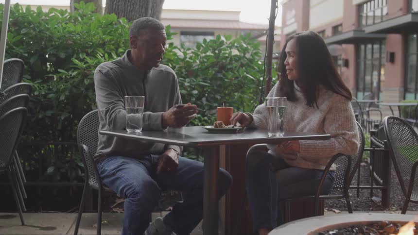 Two people sit outdoors at a cafe table engaged in conversation