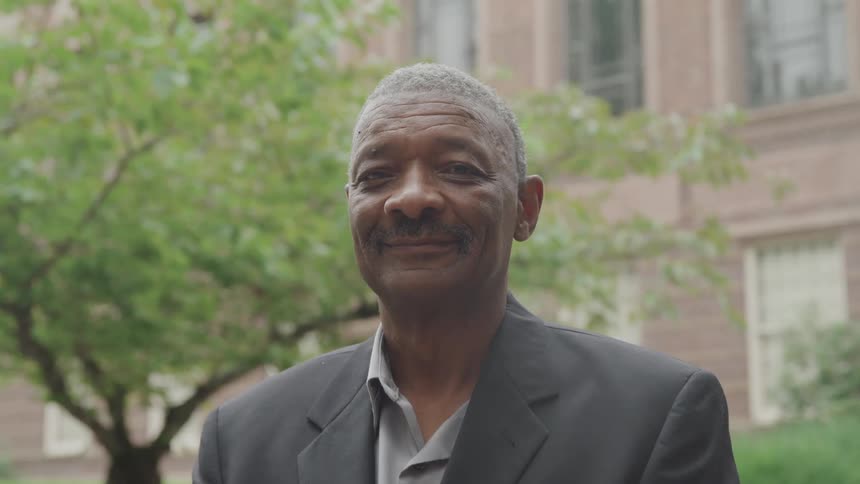 An older man smiles warmly while standing outdoors in front of a building