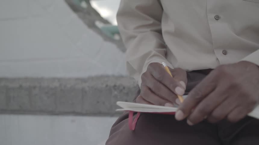 A man with dark skin and a white beard looks downward beneath a cloudy sky wearing a tan shirt and brown pants holding an open notebook and pencil as he writes or draws