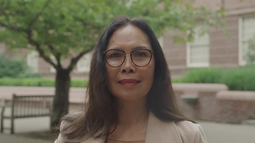 A middleaged woman with long hair wearing glasses sits outdoors in the neighborhood around midday