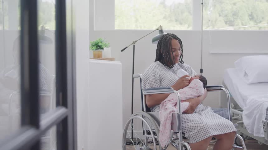 A woman in a hospital gown sits in a wheelchair bottlefeeding a baby wrapped in a pink blanket in a brightly lit room with a bed