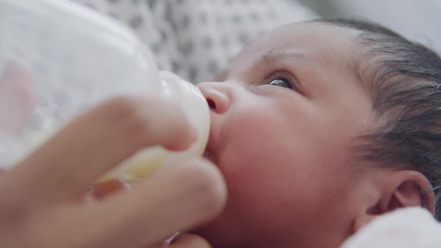 A mother feeds her infant child a milk bottle as the infant gazes up at her