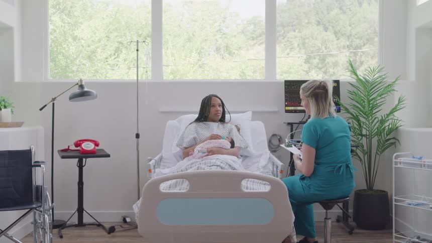 A young woman lies on a hospital bed holding her newborn child with a nurse sitting beside her and talking to her around midday