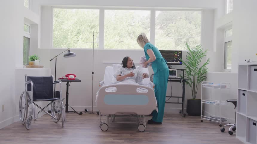 Nurse handing a newborn baby to the mother who is lying on a gurney in a hospital room