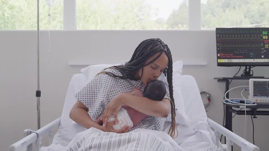 New mother sitting in a hospital bed holding and kissing her newly born baby