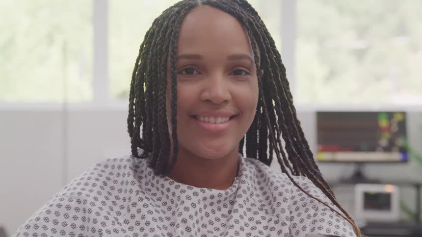 A woman with dark braids and a patterned hospital gown smiles broadly while looking at the viewer