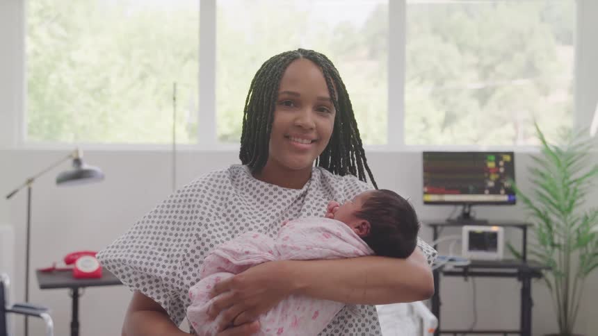 A person in a hospital gown cradles a newborn wrapped in a pink blanket surrounded by medical equipment and a window overlooking lush foliage