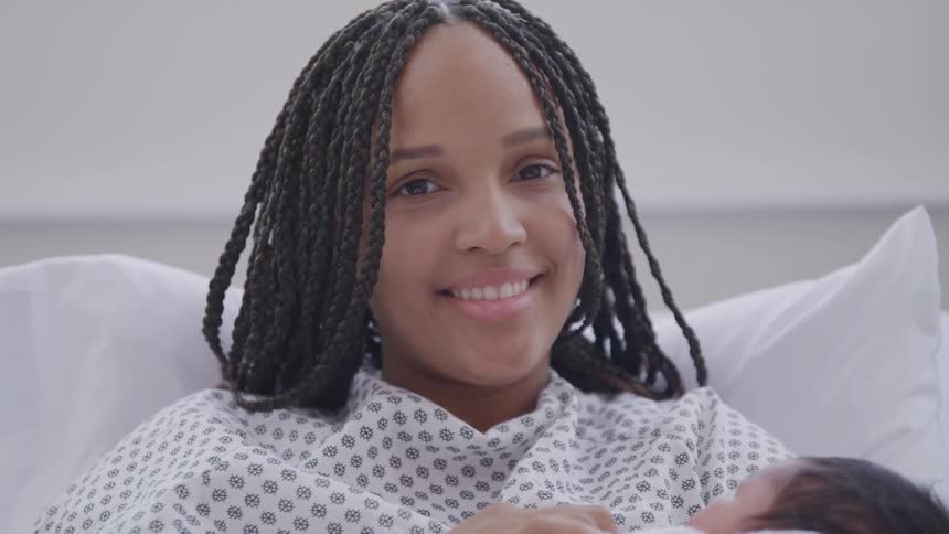 A woman with braided hair lies in a bed smiles and holds a baby