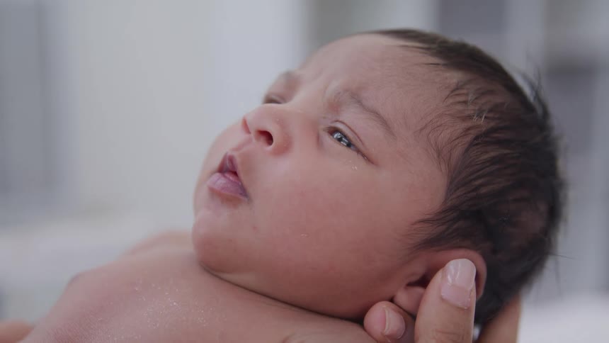 A newborn baby is gently held and gazes upward with a curious expression