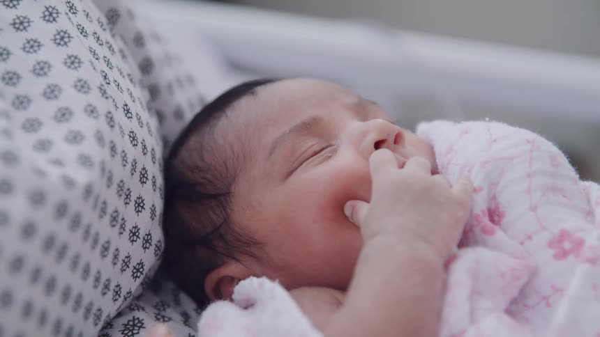 Close up of a baby sleeping in his mothers arms as she sits on the hospital bed