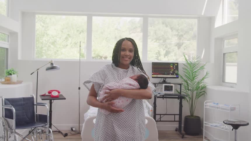 A smiling mother in a hospital gown holds her newborn baby in a bright hospital room
