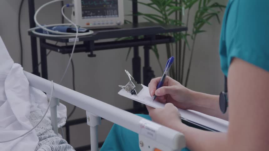 A medical professional in scrubs writes on a clipboard next to a patients bed with a vital signs monitor in the background