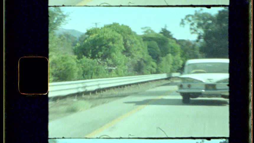 A vintage car drives along a highway surrounded by trees seen through an old film frame